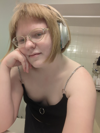 Many, a white queer in a bisexual as hell bob haircut with straight-cut bangs (and a little cowlick, lol), is leaning over. Fae're wearing a low-cut Hot Topic strappy top, where the straps are chains attached by o-rings. There's a lot of cleavage going on. Fae've also got cute glasses on. Adorable overall.