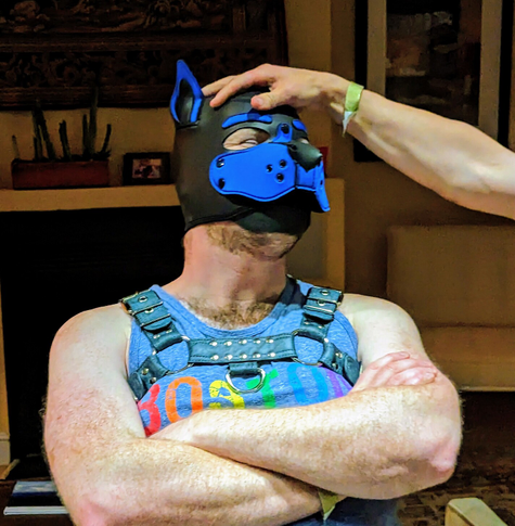 Crowbar with crossed arms in pup hood, tank top, and harness, receiving head pat from a friend