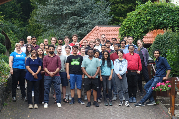A group of about 40 people, mostly young and with diverse appearances, posing for a picture on a paved slope in front of huts and a forest. They are looking towards the camera and are mostly smiling.
