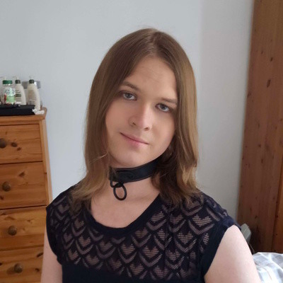 A transfeminine non-binary woman sat on a bed, and wearing a collar with a lacey top. She has a slight smile and looks into the camera.