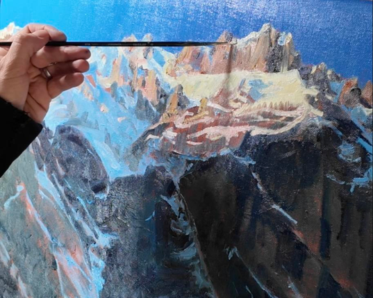 Die linke Hand der Künstlerin hält einen feinen Pinsel und malt an einem mittelgrossen Bild (ca. 50 x 70 cm). Das Bild, welches gerade entsteht, ist von einem realistischen, felsigen, schneebedeckten Berggrat. Die letzten Sonnenstrahlen berühren eben noch die obersten Felsen und Schneeflächen und tauchen es in ein warmes, goldenes Licht. Der Himmel darüber ist tief blau. Die unteren 2/3 des Berges liegen bereits im dunklen Schatten. Das noch unfertige Bild drückt bereits eine tiefe Ruhe aus.