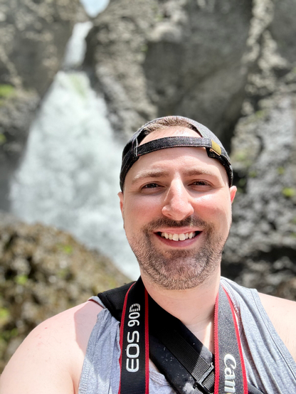 Scruffy man in a tank top and backwards baseball cap smiling in front of a waterfall.