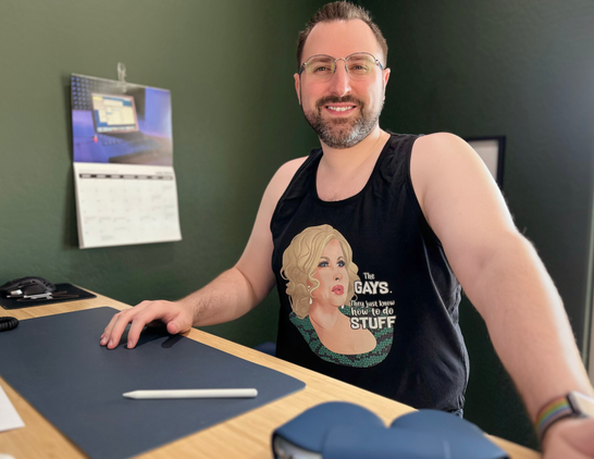 Smiling man with beard and glasses. The glasses are round, half-frameless, with blue accents. Wearing a black tank top with a caricature of Jennifer Coolidge from the movie “Single All The Way” and the quote “They gays. They just know how to do stuff.