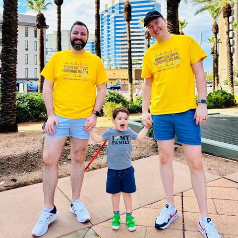 "A toddler standing between his dads at a pride parade on a sunny day.