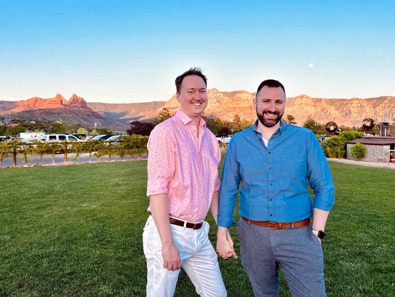 Two men smiling and holding hands at sunset with Sedona, Arizona in the background.