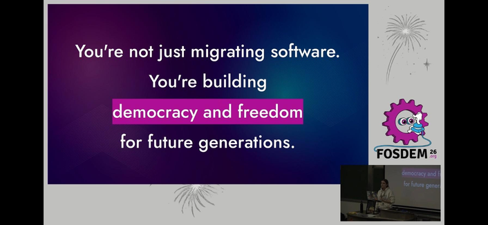 The image features a slide that reads: "You’re not just migrating software. You’re building democracy and freedom for future generations." It has a colorful background and displays a logo for FOSDEM 2026, accompanied by a speaker at a podium.