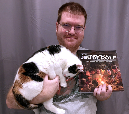 Julien Pirou souriant devant un fond gris, tenant un chat d'une main et le livre La Grande Aventure du Jeu de Rôle de l'autre.