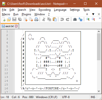 An image of ASCII art from within a text editor. The art is the head of my fursona Foxfire, an anthro fox, smiling and looking at the viewer. His fangs are slightly visible under his muzzle, and he has blush marks on either side of his face. A shine shape is on the top left, above his head. Foxfire is on the top face of a cube, with the two other visible faces on the left and middle being filled with text to shade them. Foxfire's name is seen on the bottom middle face. 
