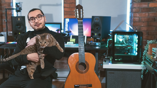 Adrian sitting wearing all black in a turtle neck holding his cat, music studio with exposed brick walls and computer workstation in background, nylon string guitar propped up in center