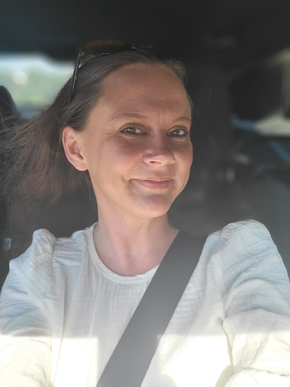 Selfie of melsaywhat as a passenger in a car with her seatbelt on. Sunglasses atop her beginning to gray on top brown hair, smiling. Wearing an off-white long sleeved crew neck blouse with slightly ruched shoulders at the sleeve divide. Also, I'm horrible at alt text, but I'm working on it.