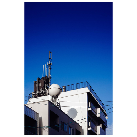A mid-rise apartment building photographed from below against a deep blue sky. On the rooftop, a cluster of cell antennas rises beside a white spherical tank mounted on metal supports. The sphere casts a sharp circular shadow onto the pale exterior wall. Dark power lines run diagonally across the frame, and stacked balconies with black railings line the right side of the building.