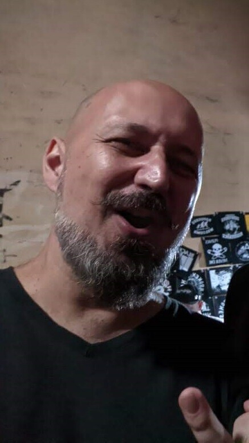 Selfie: light skinned latino man in his forties, bald, skinny, with a big nose. With a short gray beard, pointy mustache.  I'm making heavy metal horns with my fingers and saying "whoo hoo" at a rock concert.  Wearing a black T-shirt, after all, it was a rock'n'roll concert and it is mandatory by law to wear black. 