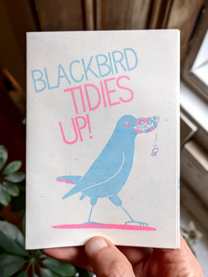 Photo of a  white dude's hand (who might it be!) holding a small zine. On the cover is a giant blackbird in pink and blue, a car in it's beak, it says „Blackbird Tidies Up!“ next to it.
