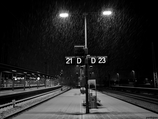 Early in the morning, snow covers a quiet platform where illuminated signs displaying the numbers 21 D and 23 D can be seen, and the falling snow sparkles in the light.

Am fruehen Morgen bedeckt Schnee einen ruhigen Bahnsteig, auf dem beleuchtete Schilder mit den Zahlen 21 D und 23 D zu sehen sind, und der fallende Schnee funkelt im Licht.