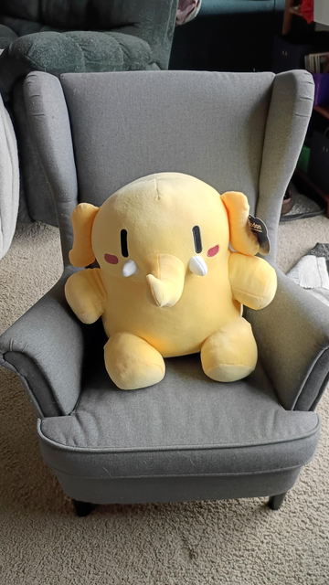 a small size chair with the large tan mastodon plushie, which is a cartoon elephant shaped critter with vertical dot eyes and oval blushes cheeks.  finally a cute trunk with tiny tusks.  the plushie comfortably fills the chair.