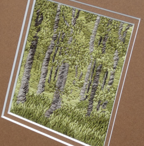 Stitched artwork depicting a summer woodland with light and shade. A very breen pallette with tall thin tree trunks in brown/grey. Close mounted in a brown double mount. Viewed on an angle to show texture.