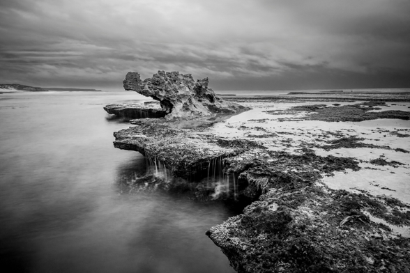 Black and white landscape of dragon's head rock. This long exposure features a moody cloudy sky and water running off the rock formation to the ocean just underneath. 