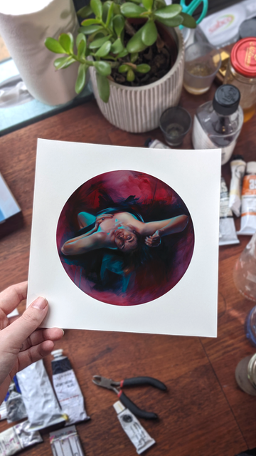 a hand holds an art print over a busy artists table strewn with art supplies, tubes of paint, a plant and jars of medium. the print is a tondo of a woman leaning back and looking forward, her face stressed from the pull of gravity in the deep bend