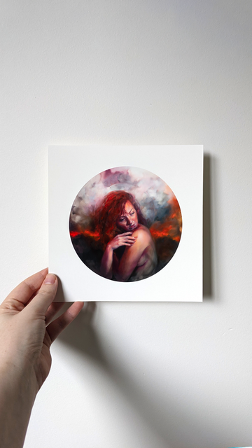 a hand holds an artwork print against a white wall, the print is a tondo painting of a woman holding herself in greif, her red hair thrown to one side. the background is an abstract horizon of fire and smoke clouds