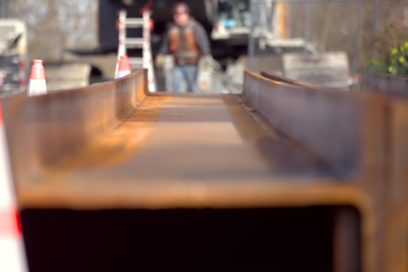 a low, artistic angle shot above a slightly rusted I beam laying toward the camera long-wise. Blurred in the background can be seen construction equipment, traffic cones, and a construction worker in personal protective equipment