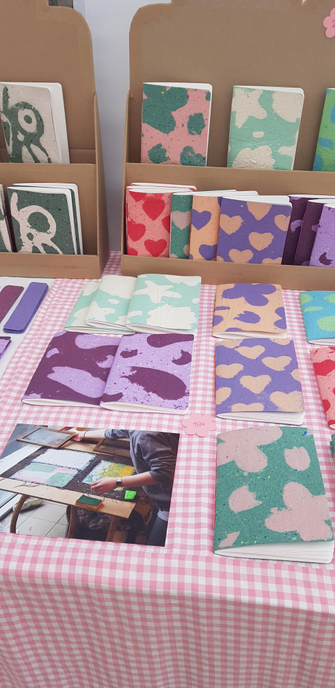 Gros plan de mon stand sur les marchés avec des carnets en papier recyclé. Il y a des couvertures avec des motifs avec des formes de fleurs, de cœurs, de coquillages et de papillons.