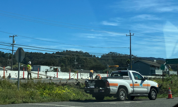 A CalTrans truck parked along a frontage road; closer to the highway but protected by large white barriers, CalTrans folks are working on a new bus stop. It's a sunny day with a bright blue sky. 