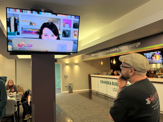 A man stands in front of a television displaying a close-up of a woman working on a laptop. The setting is a modern lounge area with a bar and seating, featuring a sign for the Swansea Building Society Lounge.