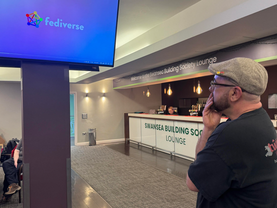 A man stands with his hand on his chin, looking at a large screen displaying the logo "fediverse."