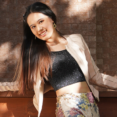 Photograph of Tanya Kan, a smiling woman with long black and brown hair, sparkly black crop top, and pink pleather jacket. She is standing and leaning against a railing outdoors.