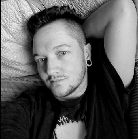 Black and white pic of a 46 year old trans man with dark hair and facial hair.bhe reclines with one arm above his head, and looks at you with a slight smile.