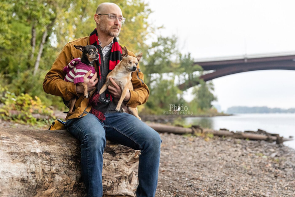 A white, middle-aged, bald, bespectacled man with a beard. He's sitting on a river bank log with a bridge in the background to the right. The the left are autumn trees. In his lap are two small, adorable dogs, one blonde, one black and tan. Everyone is looking off to the right.