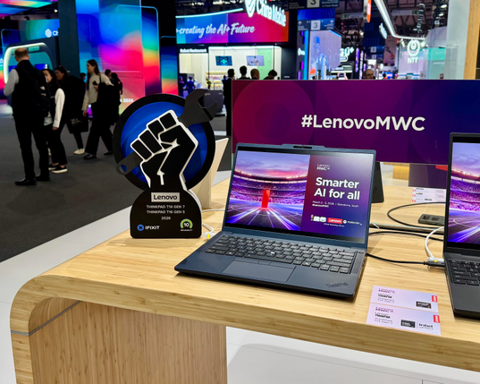 A Lenovo ThinkPad laptop sits on display at a trade show booth beside an iFixit repairability award trophy shaped like a raised fist holding a wrench. A sign behind the table reads “#LenovoMWC,” while colorful expo booths and attendees fill the background.