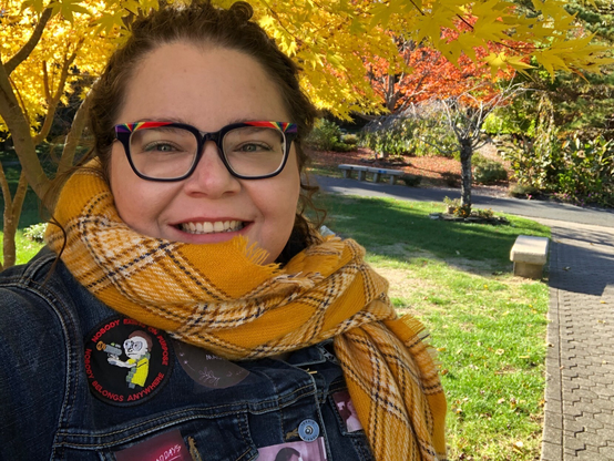 Photograph of smiling white woman with brown hair, rainbow glasses, yellow scarf, and denim jacket with patches in front of yellow autumn trees.