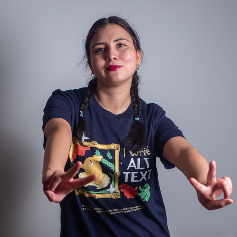 A beautiful woman with twin braids wearing a navy colored t-shirt featuring the slogan "I write alt text" next to a cartoon reproduction of the Birth of Venus, with Venus portrayed by a cute elephant. Next to the painting, there are two other elephants, one of who is wearing dark glasses and is holding a cane, while the other is guiding him and describing the painting. Underneath, there is a textual description of the above design.