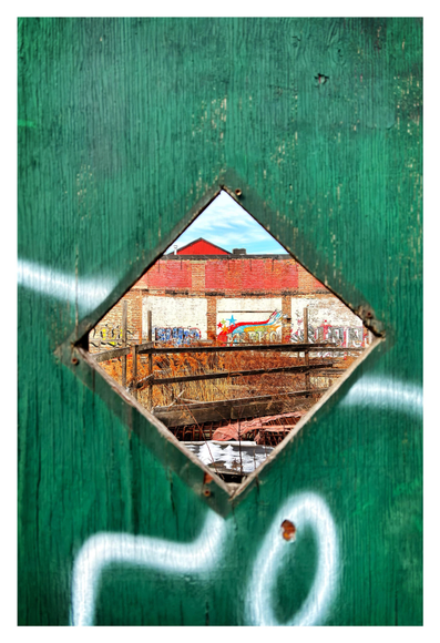 The view through a diamond-shaped cutout in a plywood barrier around a vacant city lot. The textured surface of the plywood is painted green with some wavy lines and loops of white spray paint. Through the cutout we can see assorted odds and ends: some decaying wooden railings, a rumpled piece of orange construction netting, a black tarp underneath clumps of melting snow. Tall brown reeds or grasses push up in all the in-between spaces. In the background is a brick wall with some graffiti in vibrant colors. Beyond the wall is a glimpse of a red triangular rooftop, under a triangle of clouds and blue sky.