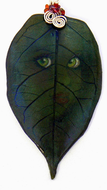 Handmade polymer clay pendant shaped like a dark green leaf with delicate veins and a mysterious nature spirits  face subtly emerging from the depths. Garnets and fire opals  are woven with sterling silver wire into two larger spirals, giving the piece a magical forest spirit aesthetic.