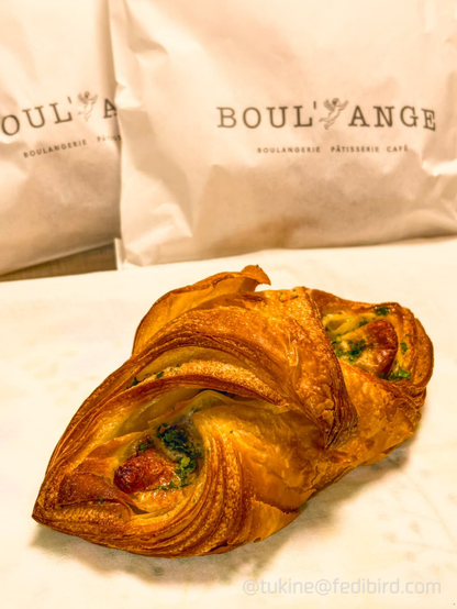 BOUL'ANGE（ブールアンジュ）の「笹かまデニッシュ」。何層にも重なった黄金色の香ばしいデニッシュ生地の間に、焼き色のついた厚切りの笹かまぼこと、刻んだ大葉（シソ）が包まれています。背景には店名のロゴが入った白い紙袋が写っています。