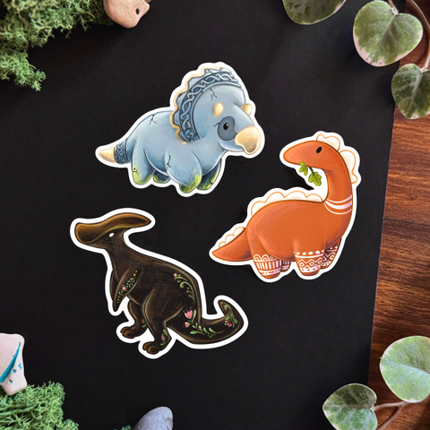 Picture of 3 stickers set on a black and wooden surface framed with decorative plants and stones. 
The three stickers each represent a different dinosaur. The top one is a triceratops that seems to be made of stone carved with Celtic designs. Its horns, beak, and tail tips are bone-coloured. On its legs, moss is slowly growing. 
The middle one is a Terra-cota coloured diplodocus munching on a leaf. Geometric white patterns, similar to those painted on terra cota vases, decorate its four legs, and two stripes decorate its neck. The rounded spikes on its head and back are white. 
The last dinosaur is a dark brown Parasaurolophus with folk-like floral patterns painted on its tail, back and legs. 