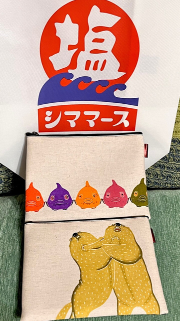 生成り色のキャンバス地のポーチが2つ縦に並んでいる。手前のポーチには、2頭の黄色いマーモットが立ち上がり、お互いの肩に手を置いてケンカをしているようなイラストが描かれている。奥のポーチには、オレンジ、紫、ピンク色などの5匹のカラフルで丸っこいダンゴウオが横一列に並んでいるイラストが描かれている。ポーチの背景には、赤、白、青で構成された「シママース」（沖縄の塩ブランド）の大きなロゴが入った袋が置かれている。