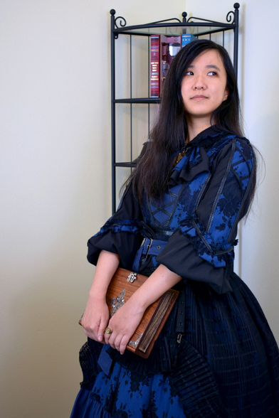 Photo: Ayu carrying a book-shaped wooden box, and dressed in a black and navy lace dress.