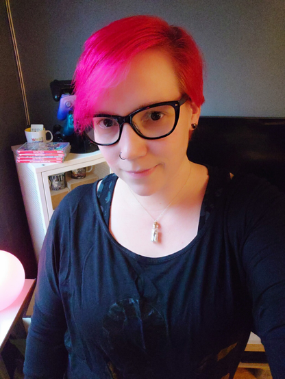 Selfie of a Rini, white cis woman, with pink short hair and black rimmed glasses in a black long sleeve shirt with a D&D logo.