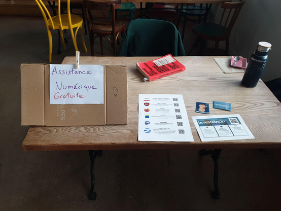 Picture of a wooden table, with a sign that says (in French) "Free Digital Support". On the table, there is a printed version of a list of apps and addons I recommend to block advertisement or as alternatives to toxic capitalist tech: uBlock Origin, Firefox, F-droid, Mastodon, Zorin OS, inventaire.io.