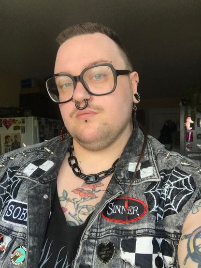 A selfie of A from the chest up, who is a white person with a brown buzzcut mohawk and braided rat tail. They have a light brown moustache and short stubble beard. have large black framed glasses, black jewelry in their eyebrow piercings, stretched septum, nostril piercings, labret piercing and 1/2" tunnels in their ears. They are wearing a thick black chain, a grey battle vest covered in patches, pins and spikes. They have an orange flower with thorns tattoo on their chest.