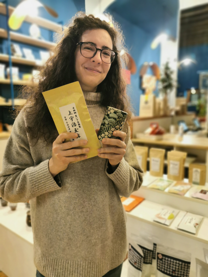 Antonella, a person with long curly hair and glasses, stands indoors in a tea shop. She is wearing a light gray turtleneck sweater and is smiling at the camera. In her hands, she holds a yellow bag of Japanese tea with Japanese characters printed on it and a small, intricately designed tea tin. The background shows shelves with various tea products and decorative items, creating a warm and inviting atmosphere.