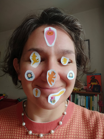 A close up of my face covoured with stickers of the main characters in different dance poses from Big Dance. I am smiling and my eyes are closed, with stickers over my eyes.