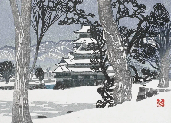 A view of Nagano's Matsumoto Castle in winter. Leafless trees stand in the foreground, screening our view of a flat snowy field stretching out toward the castle. The stacked rooftops of the multi-level castle are covered with fresh snow; the windows are dark and unlit. Beyond, low mountains cross the horizon, beneath a steely blue sky. Everything seems quiet and still.
