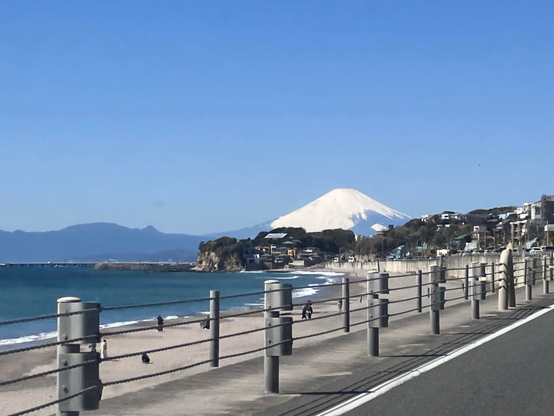 A scenic beach view featuring a coastline with people walking along the shore, a road on the right, and a snow-capped mountain in the background under a clear blue sky.