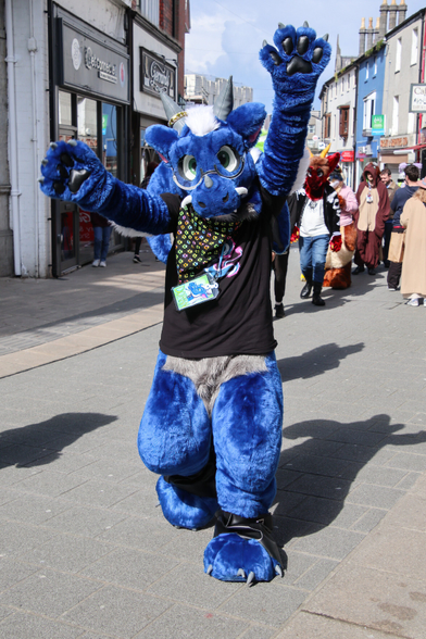A photograph of blue dragon fursuiter Doran, wearing cute round glasses and waving his hands in the air happily as he walks toward you down a pedestrianised street. Photo by Nauta Sinneau.