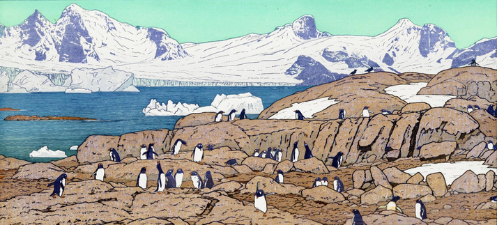 A view of a barren, rocky shoreline, somewhere in Antarctica. In the foreground, dozens of Gentoo penguins clamber over the rocks; beyond, icebergs float past, carried by calm blue water. Further on, tall jagged mountains cross the horizon, rocky peaks breaking through their snowcaps.
