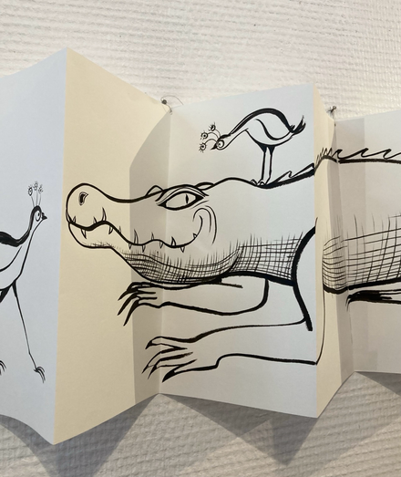A black and white drawing on a folded leperello, a long strip of paper folded as an accordion booklet, hung on a wall. It has a line drawing in black ink of a crocodile with birds on it. 
